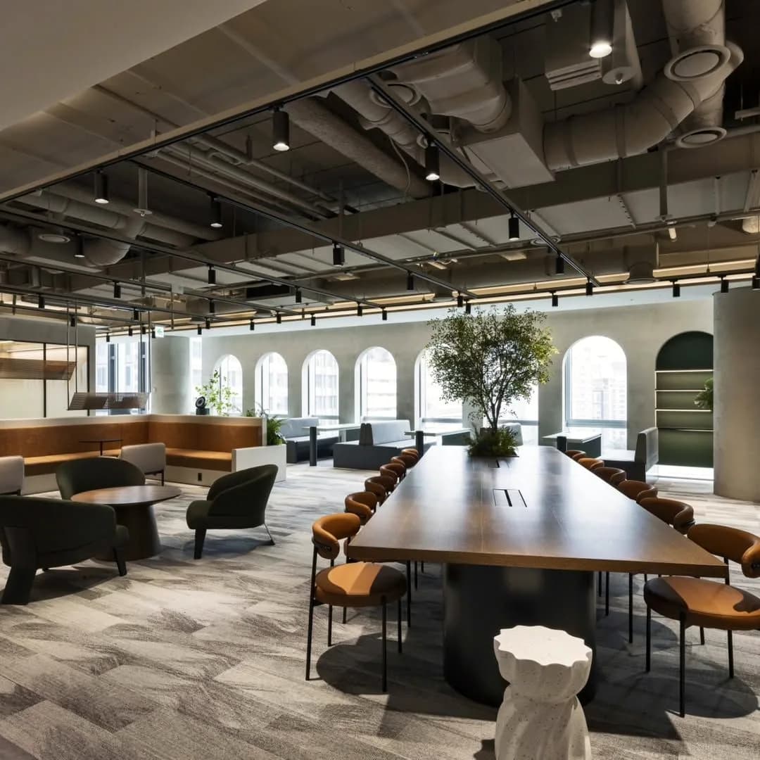 Long shared table under exposed ductwork, arched windows behind it.